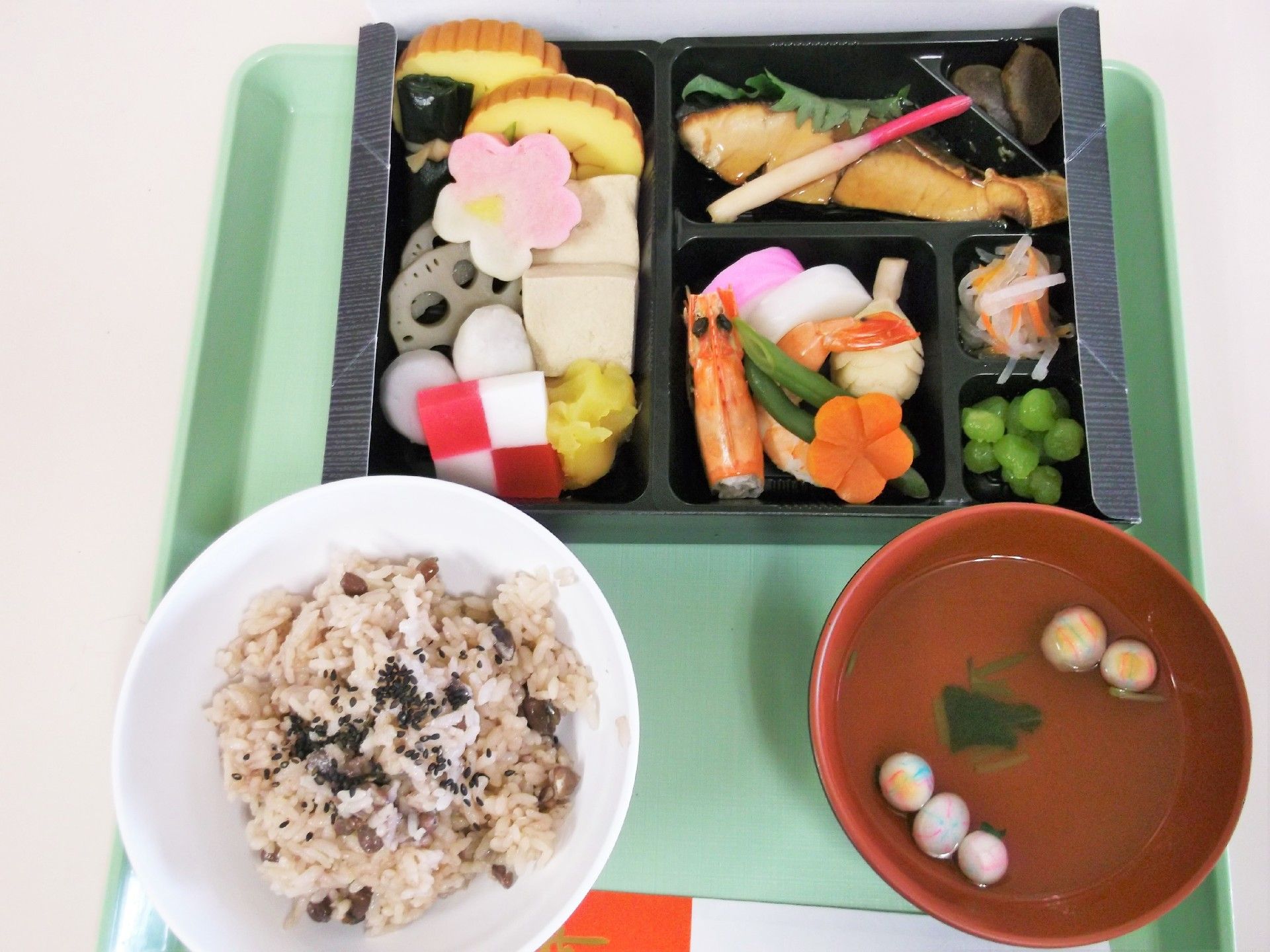 緑のトレーに並んだ赤飯に吸い物、お重風の弁当箱に詰められたごちそうの数々。