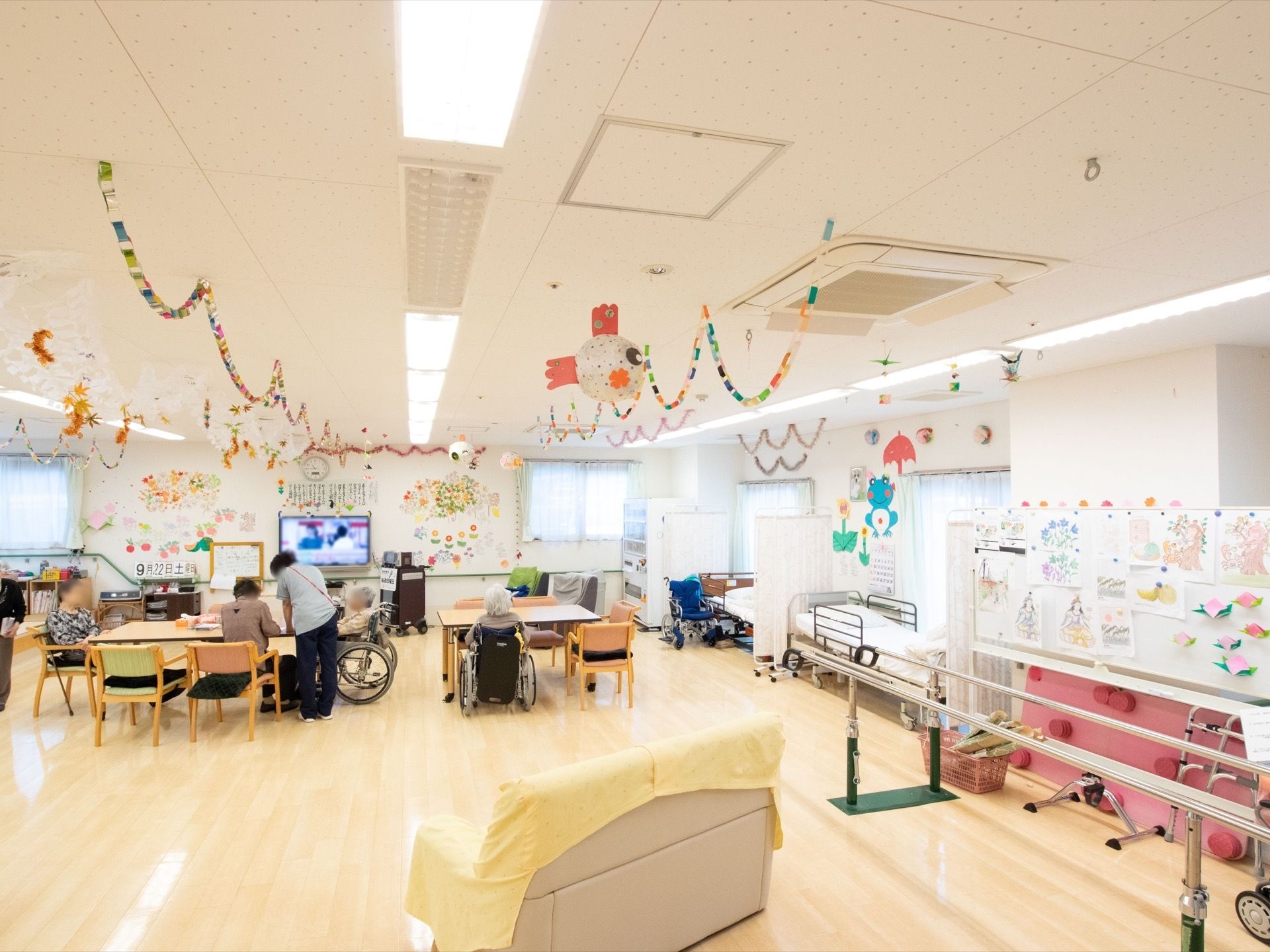 ダイニングテーブルと椅子がセットで用意されている。入居者様が数名とスタッフが一名いる。自動販売機が設けられている。
