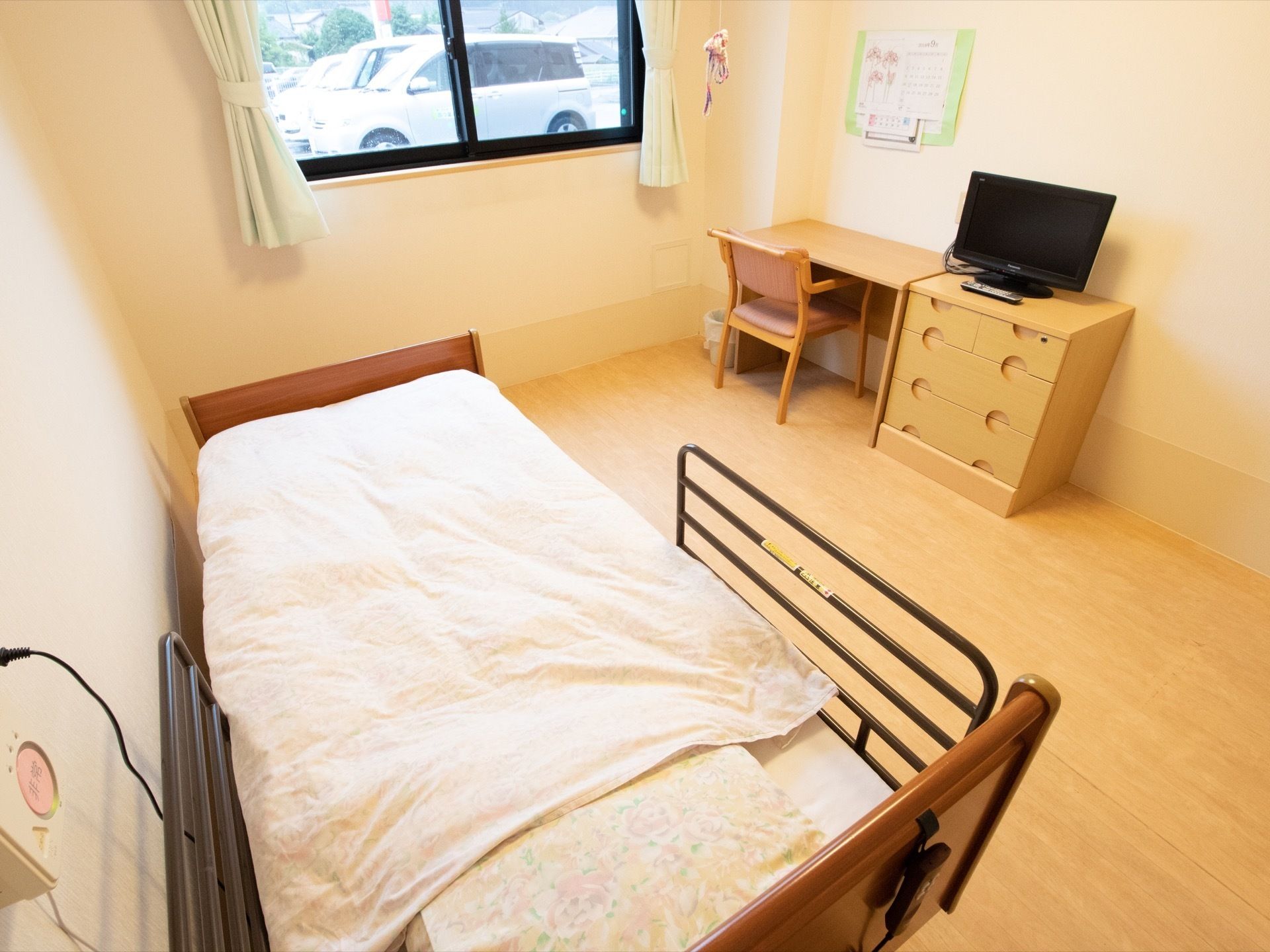 居室には窓があり、中からは外に車が止まっているのが見える。机の横にテレビが置かれたタンスがある。ベッドも用意されている。