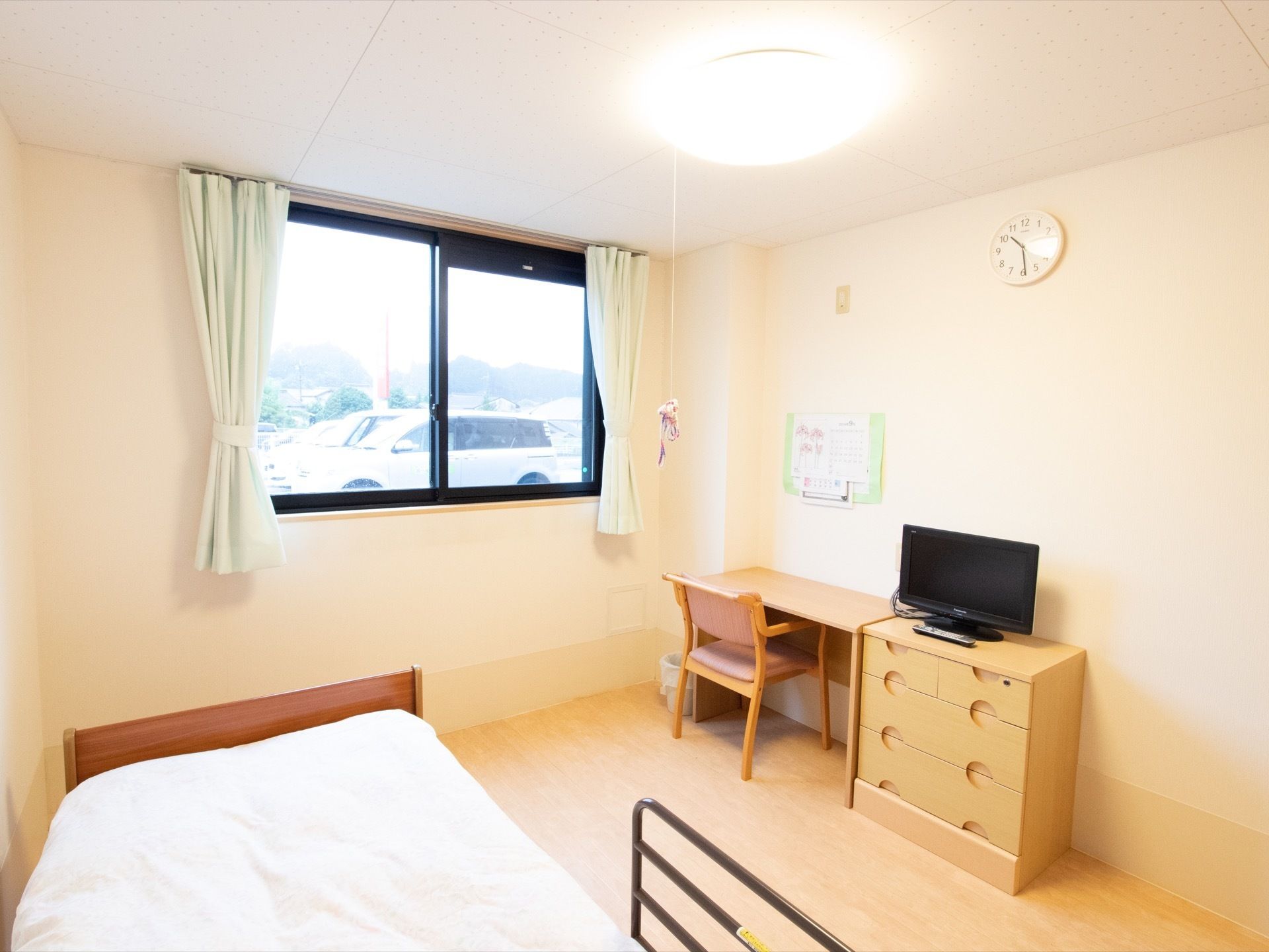 居室には机、椅子、箪笥、テレビ、ベッドが用意されている。窓にはカーテンが掛かっており、窓からは駐車場が見える。