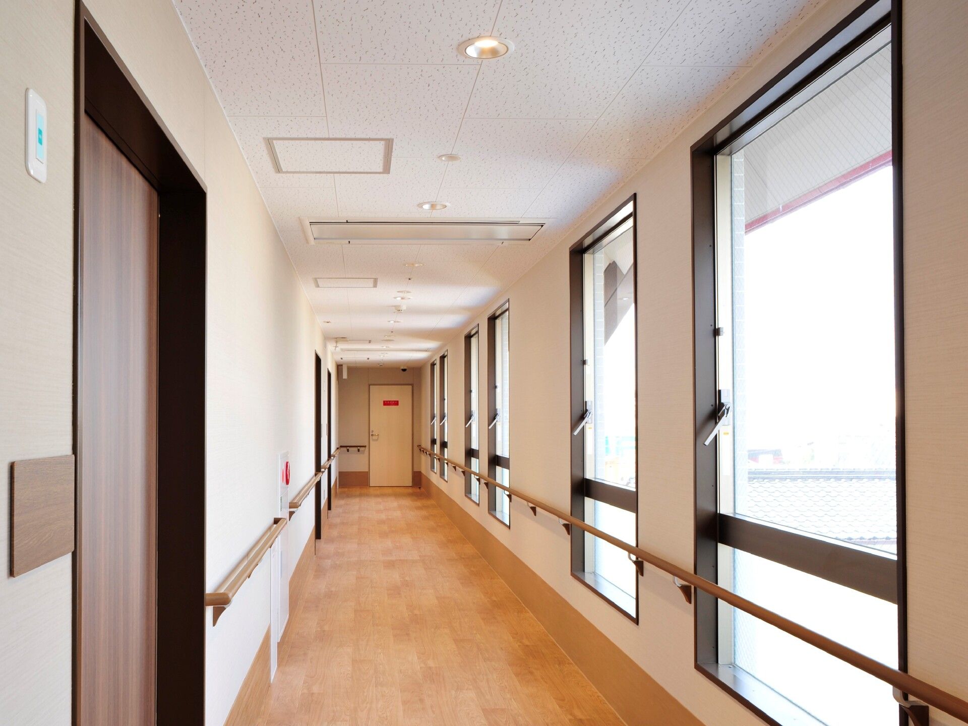 木製の手すりが両サイドに設置されたまっすぐな廊下。右側には複数の大きな窓があり、左側には居室のドアがございます