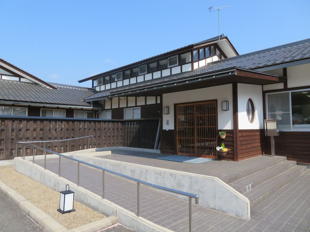 瓦屋根に白壁という純和風の建物。玄関わきには花が添えられ、階段とスロープがある。