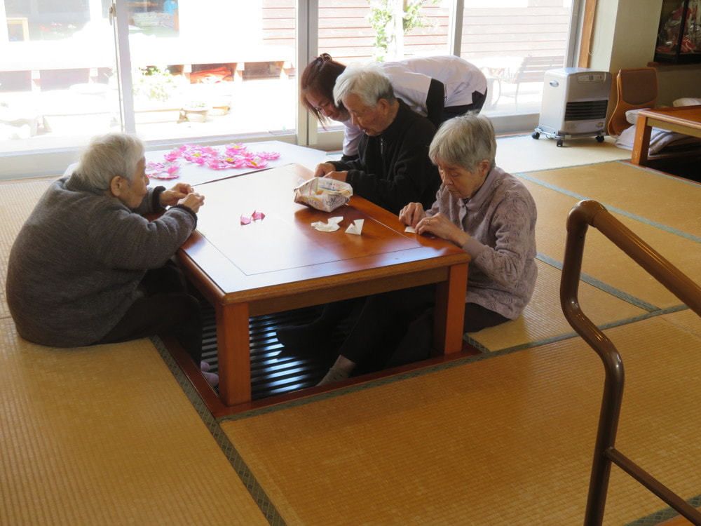 掘りごたつには3名の女性入居者がいて、ピンクと白の紙を折っている。