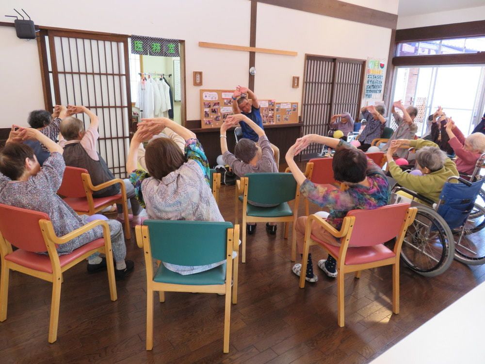 両手を頭の上で組んで伸びをしている入居者とスタッフ。車いすの入居者も参加している。