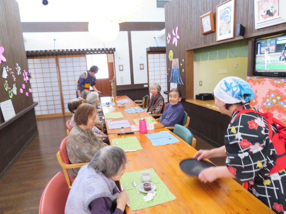 手拭いをかぶり着物を着てお茶菓子を配る女性スタッフとテーブルについている入居者たち。