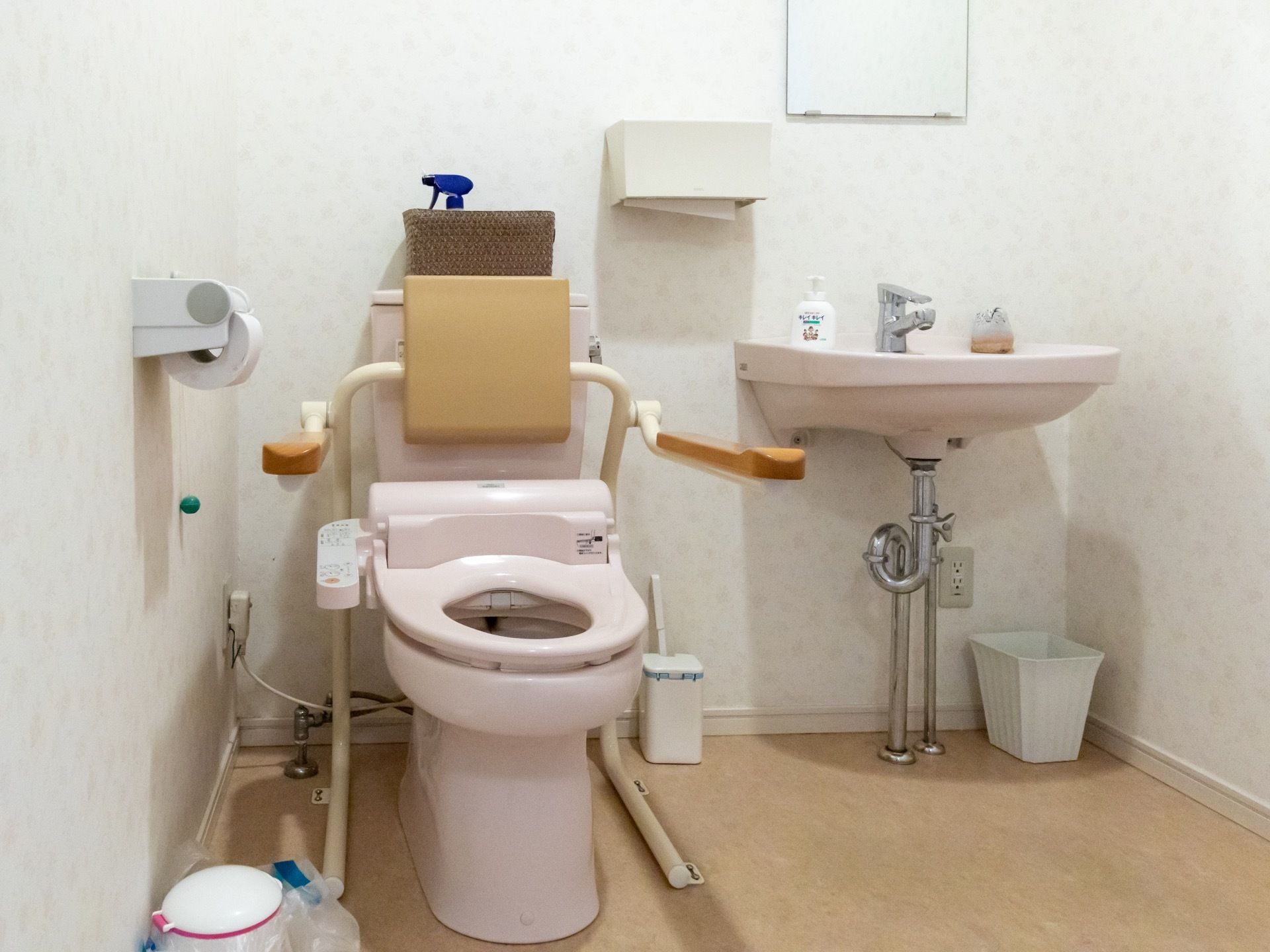 トイレの便座の左右には可動式の手すりがかかっている。タンクに濃いベージュ色の背もたれがある。