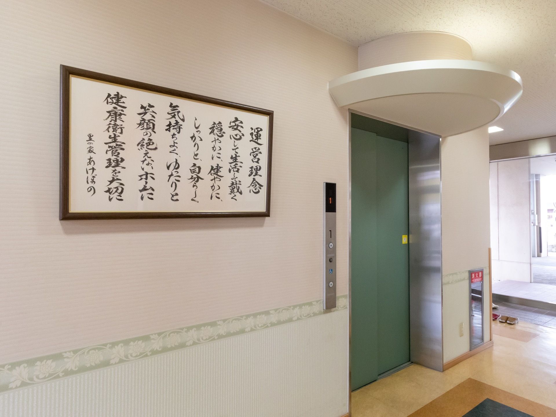 １階のエレベーターホールには、施設の運営理念を毛筆で書いた紙を額に入れて掲示している。エレベーターの近くに消火器がある。