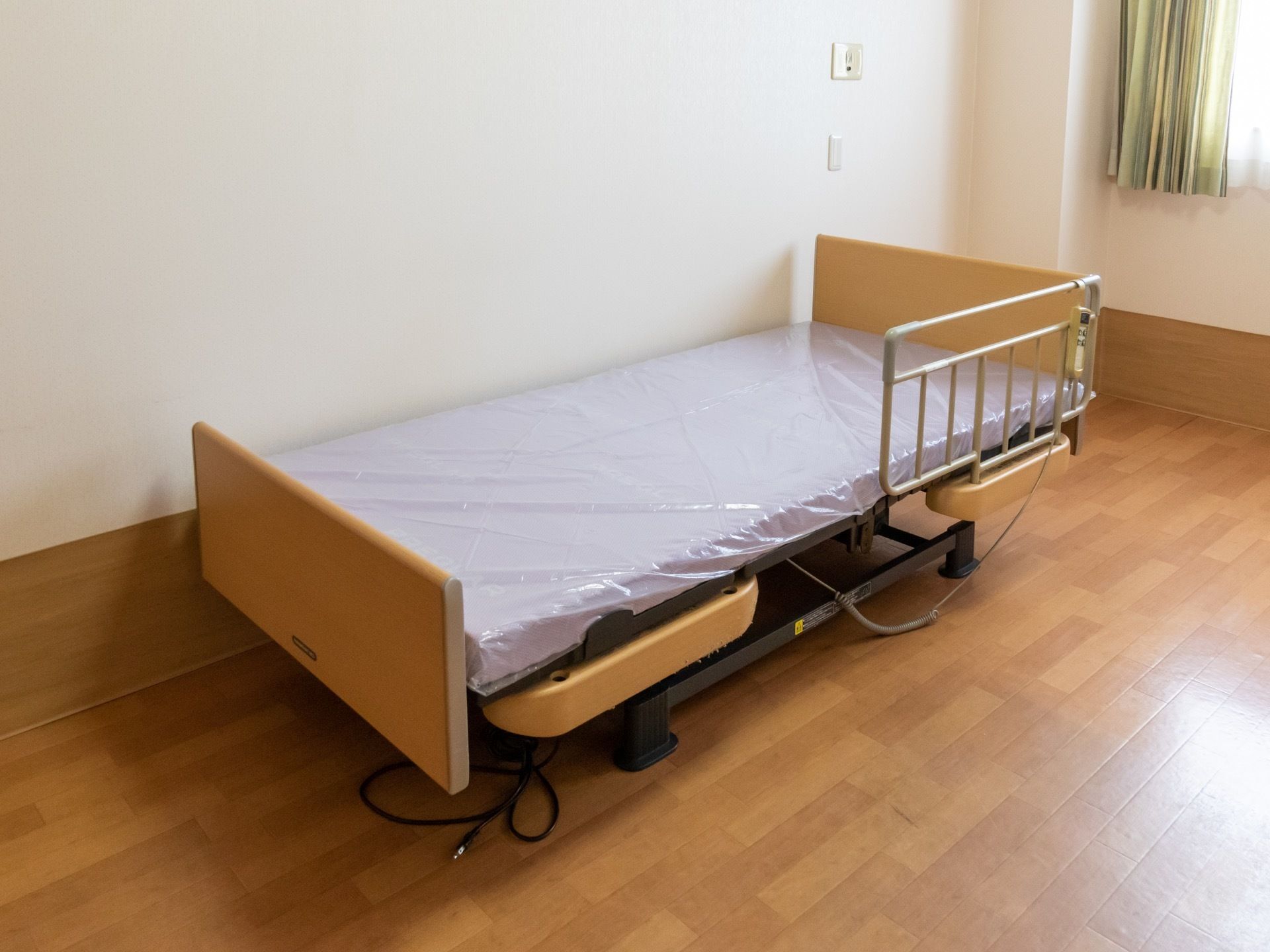 居室に導入されているベッドは電動介護用ベッドである。頭側に転落防止の柵があり、リモコンが掛けられている。