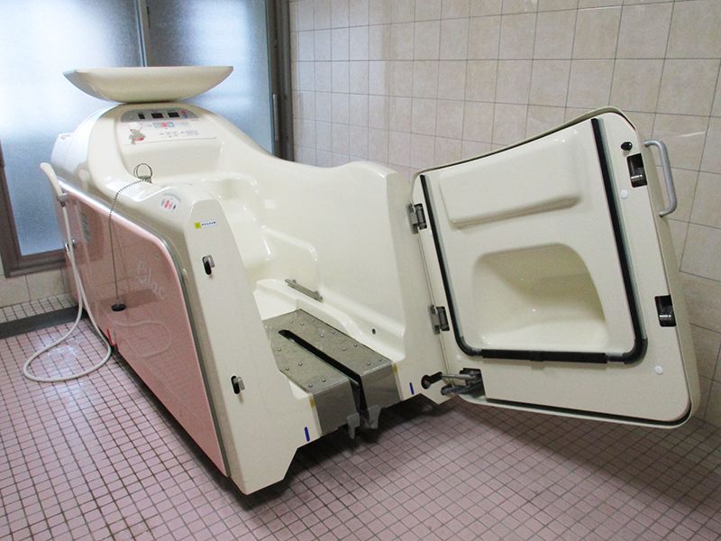 白とピンクの座ったまま入浴できる機械浴が設置されている浴室はタイル張り。