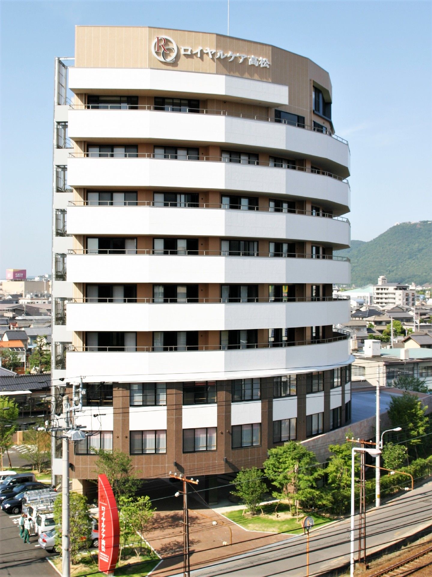 白とブラウンの重厚感のある10階建ての建物。駐車場があり緑もたくさん植えられている。