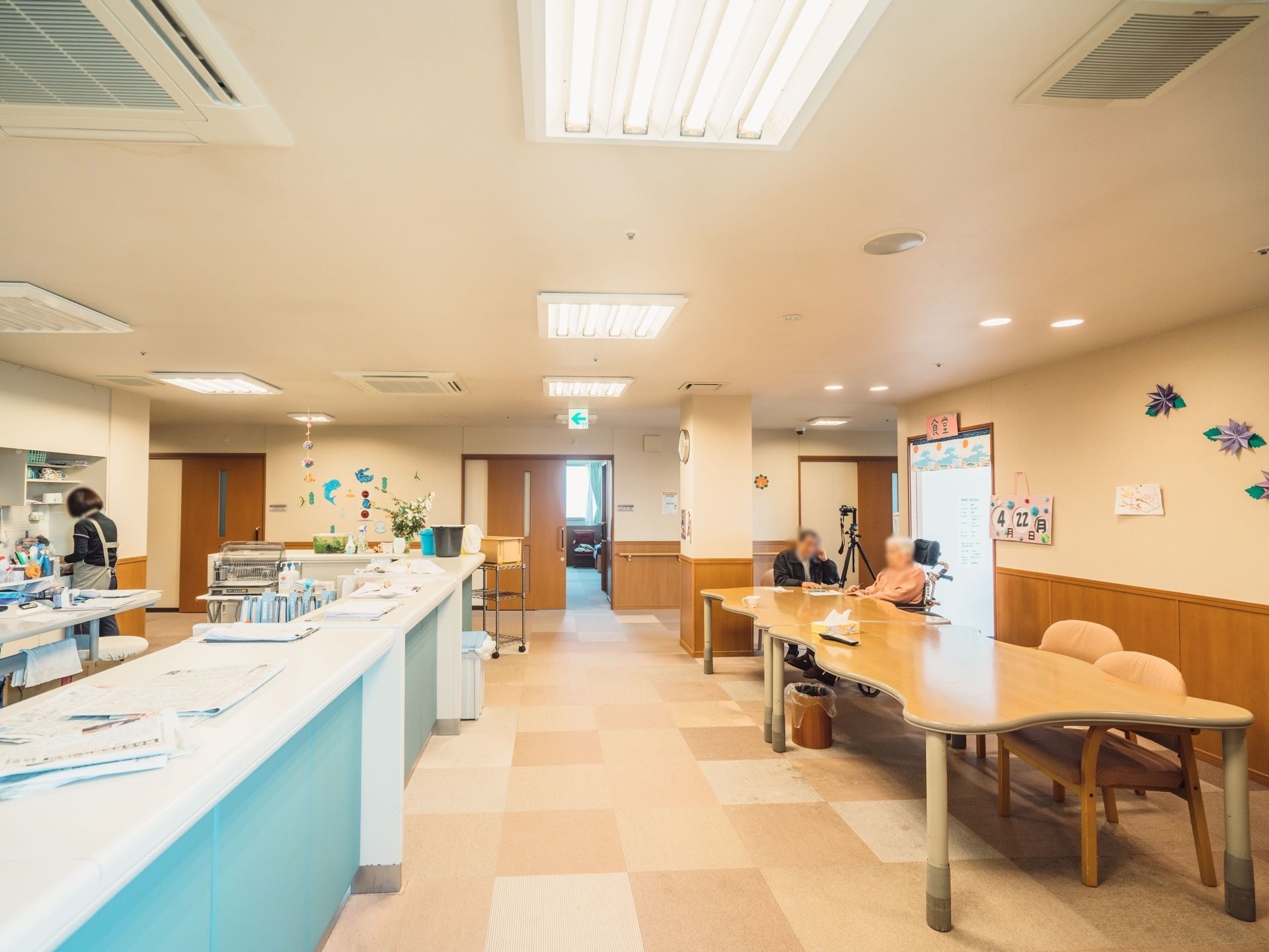 テーブルが２つ置かれ、入居者様がくつろいでいる様子である。天井には埋め込み式のエアコンが完備され、非常口への案内灯も取りつけられている。