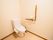 白を基調としたトイレである。壁に茶色の木製の手すりがＬ字型に設置され、床には掃除道具が置かれている。