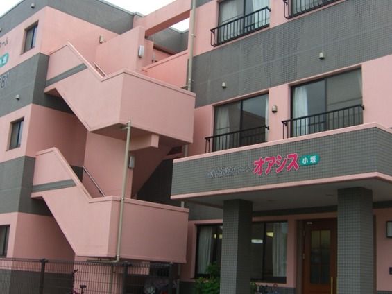 建物玄関前からアップ。玄関上の雨除け用の屋根にピンクでホーム名。ピンクとグレーの３階建ての建物。