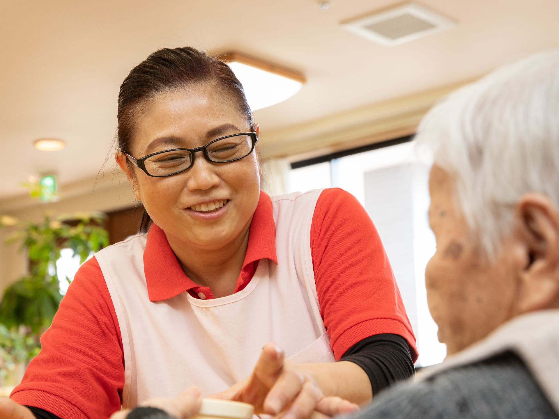 眼鏡をかけた女性スタッフが入居者様の介助を行っている。笑顔で手を取りながら、ゆっくりと話を聞いている。