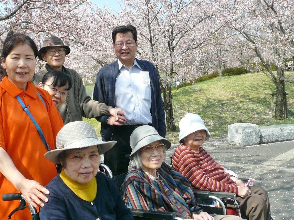 天気のいい日に桜が咲いている屋外で笑顔で過ごしている高齢者たちとポロシャツを着た女性スタッフ。