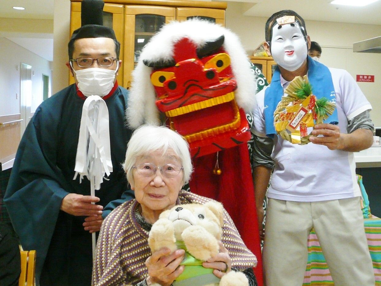 獅子舞の獅子や宮司風の男性、おたふくのお面をかぶった男性に囲まれて犬のぬいぐるみを抱いている高齢女性。