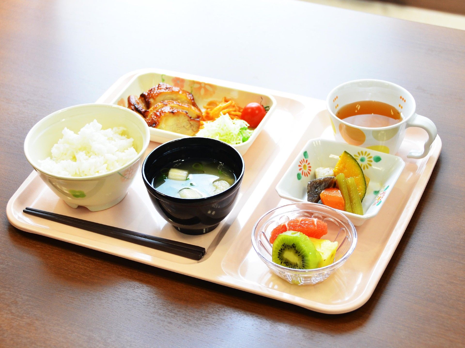 トレーの上には、ご飯、おかず、野菜、果物が彩り豊かに盛り付けられている。お茶やお箸も用意されている。