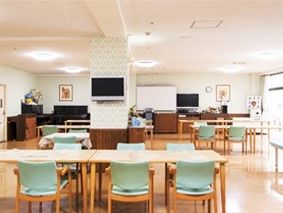 グリーンの椅子が6脚ずつセットされたテーブルが複数置かれた食堂。テレビやホワイトボードもある。