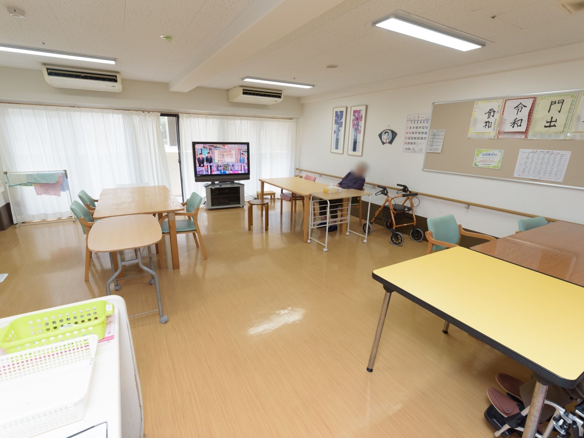 共有スペースの窓際に大型テレビが置かれている。壁には手すりがかかっている。複数のテーブル席がある。