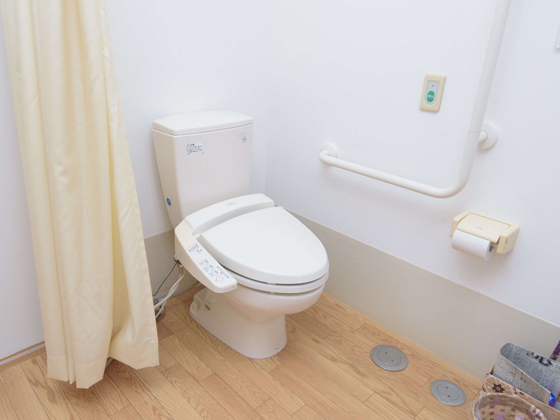 白を基調としたトイレに、温水洗浄便座が設置されている。側にはカーテンが付けられており、壁には手すりや呼び出しボタンなどが備えられている。