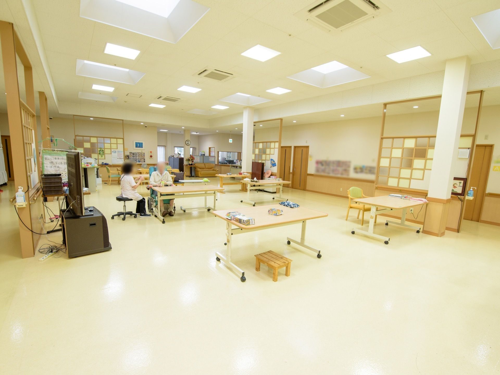 食堂は、天井が高く開放的である。テーブルが間隔を空けて設置されていて、入居者様とスタッフが椅子に座っている。