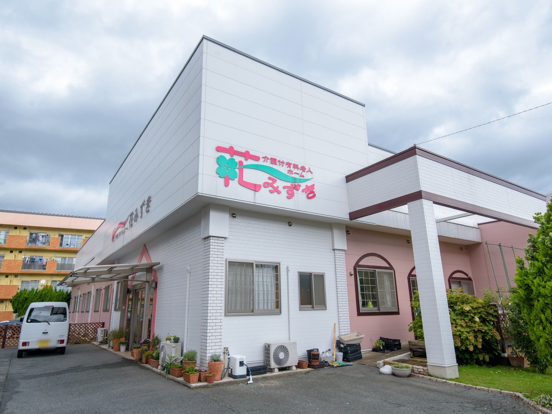 施設は白を基調とした建物で、所々の外壁がピンク色になっている。近くに、エアコンの室外機やホース、植物などが置かれている。