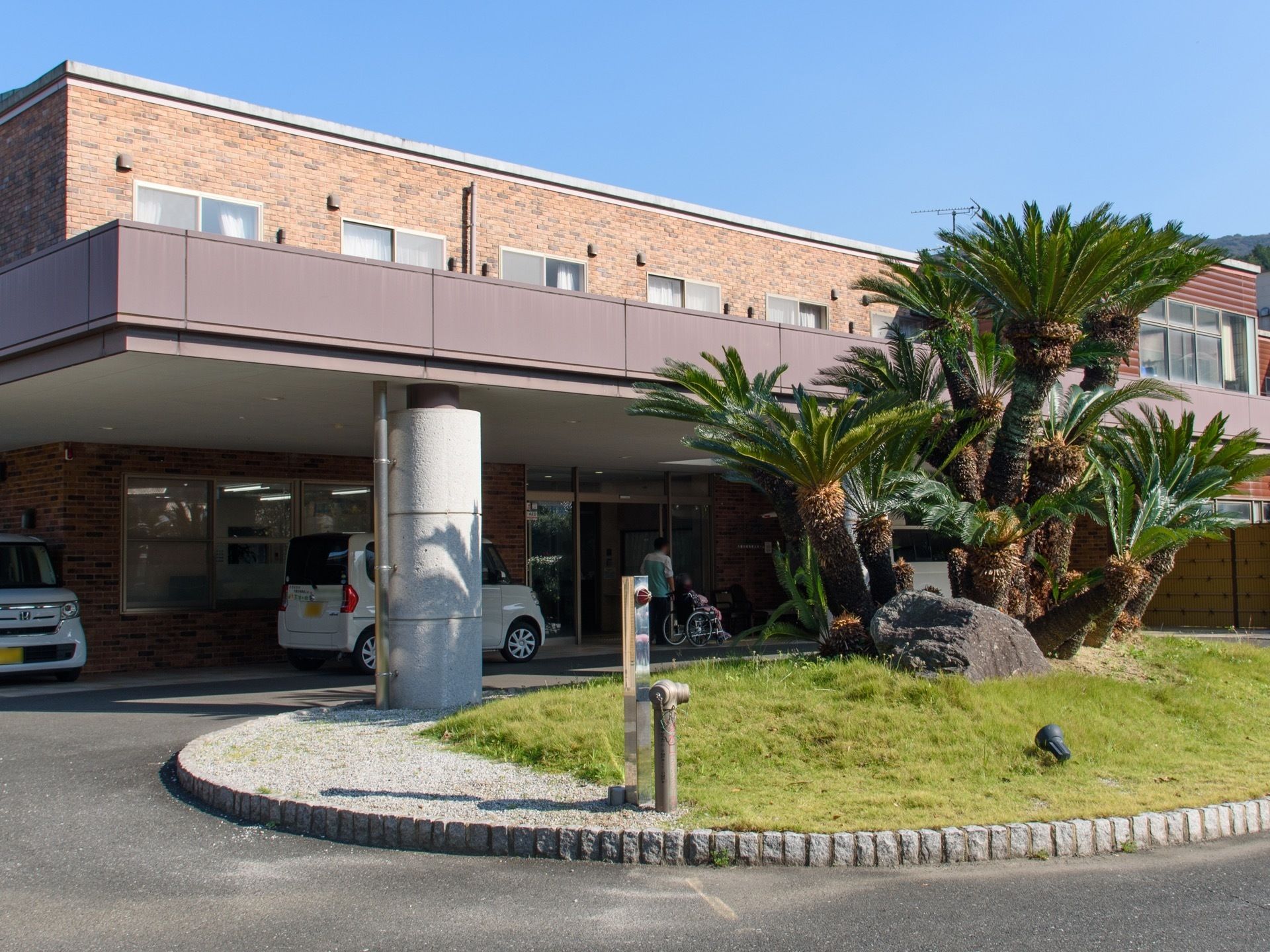外観は茶色のタイル張り。大きなひさしが設けられていて、車が乗り入れている。手前にヤシの木が植えられている。