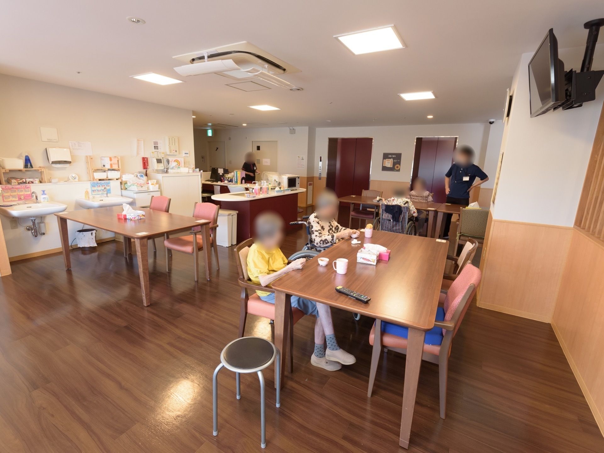 食堂には茶色い木目調のダイニングテーブルが設置されており、入居者様が席に着いている。天井にはテレビが取り付けられている。