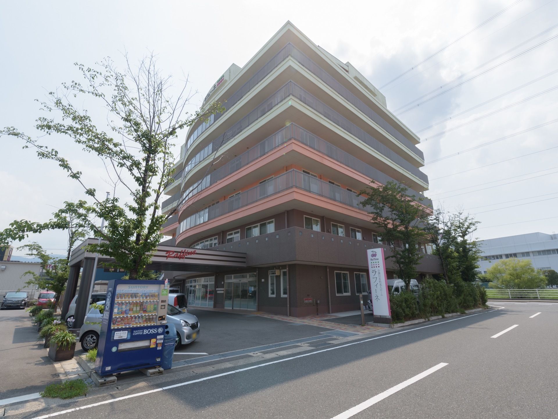 施設は茶色の外壁の建物になっており、前には道路がある。敷地の入口の右側には施設名が書かれた看板があり、左側には自動販売機が設置されている。