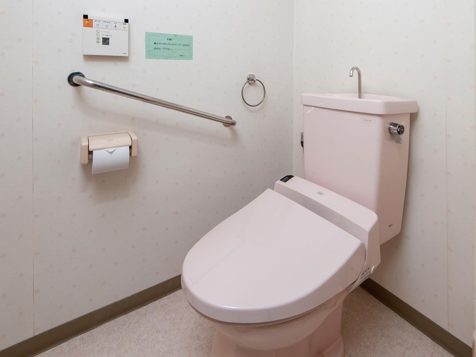 操作パネルや手すり、トイレットペーパーホルダーが壁に付いたトイレ。便座の色はピンク。
