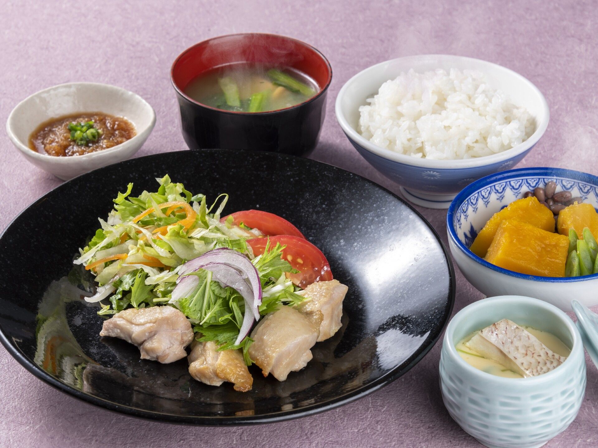 サラダを添えた鶏肉料理と茶碗蒸し、かぼちゃの煮つけ、汁物、白いご飯、小さな皿にはソースのようなものが入っている。