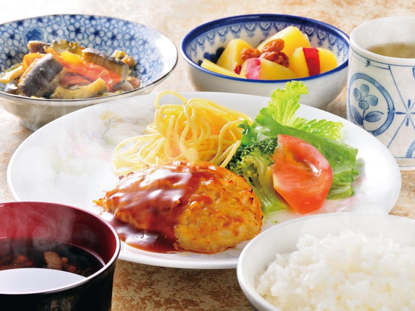 スパゲッティとサラダが添えられたコロッケのようなものに白いご飯、みそ汁、サツマイモのに煮つけ、野菜の和え物などがある。