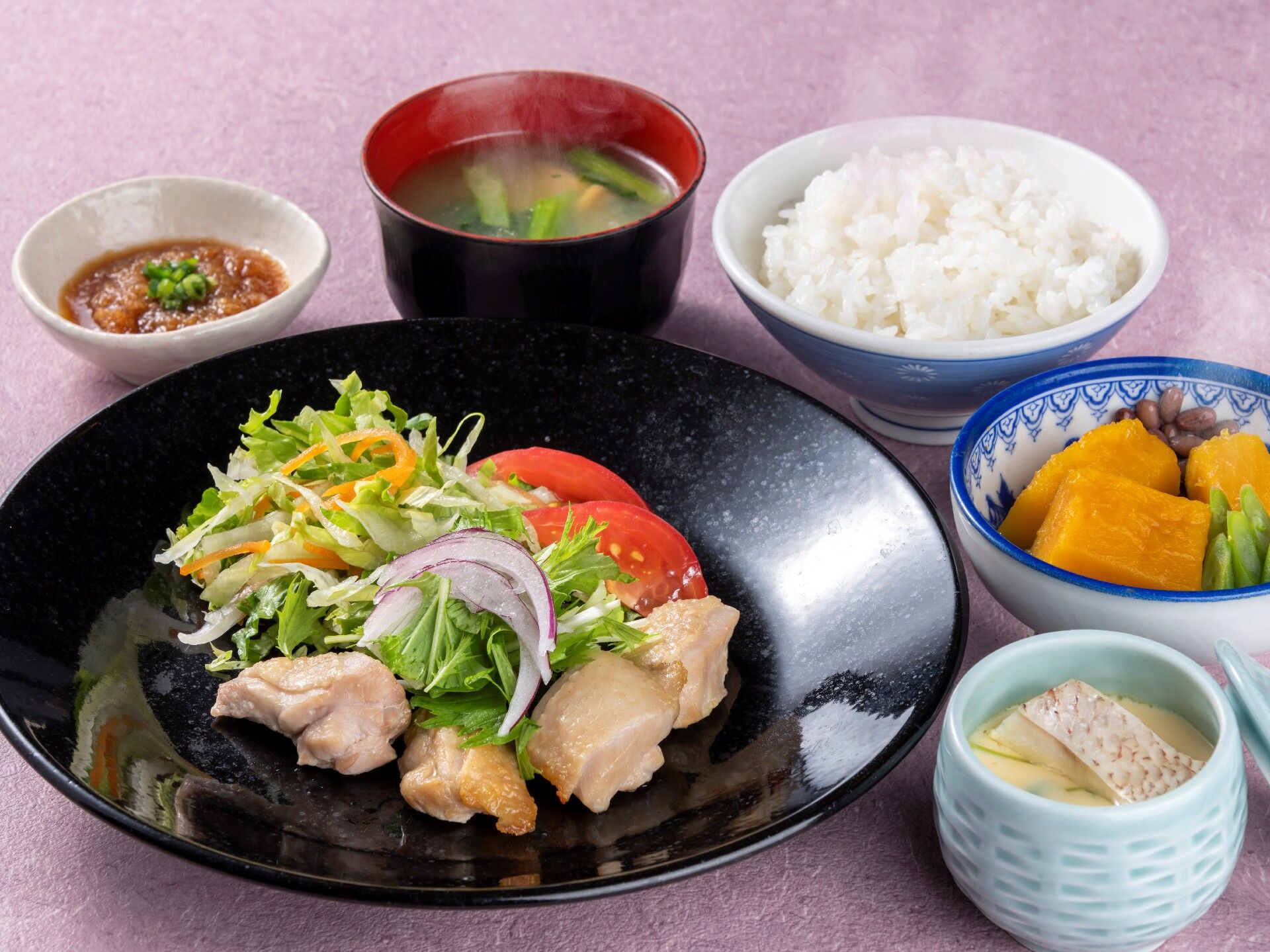 かぼちゃの煮つけや茶わん蒸し、白いご飯、みそ汁、鶏肉料理にサラダが添えられたものが並んでいる。