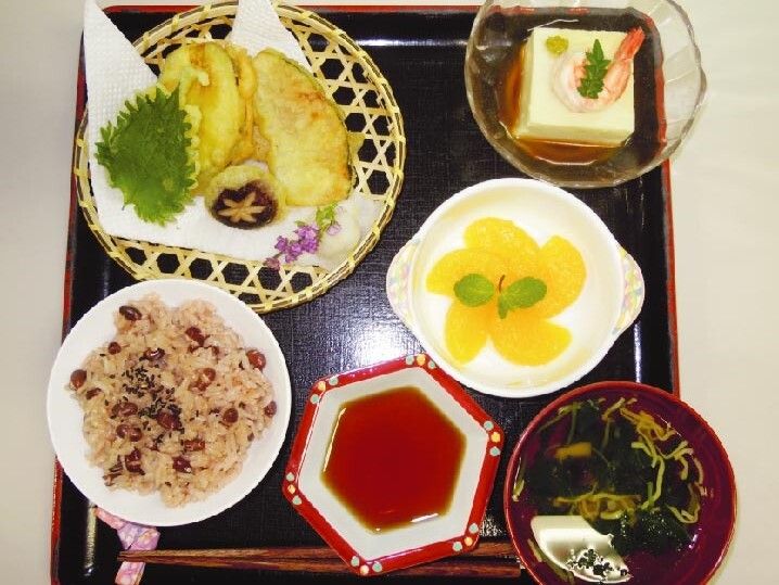 赤飯や天ぷら、汁物にフルーツが盛られた皿などが並んでいる黒いトレー。