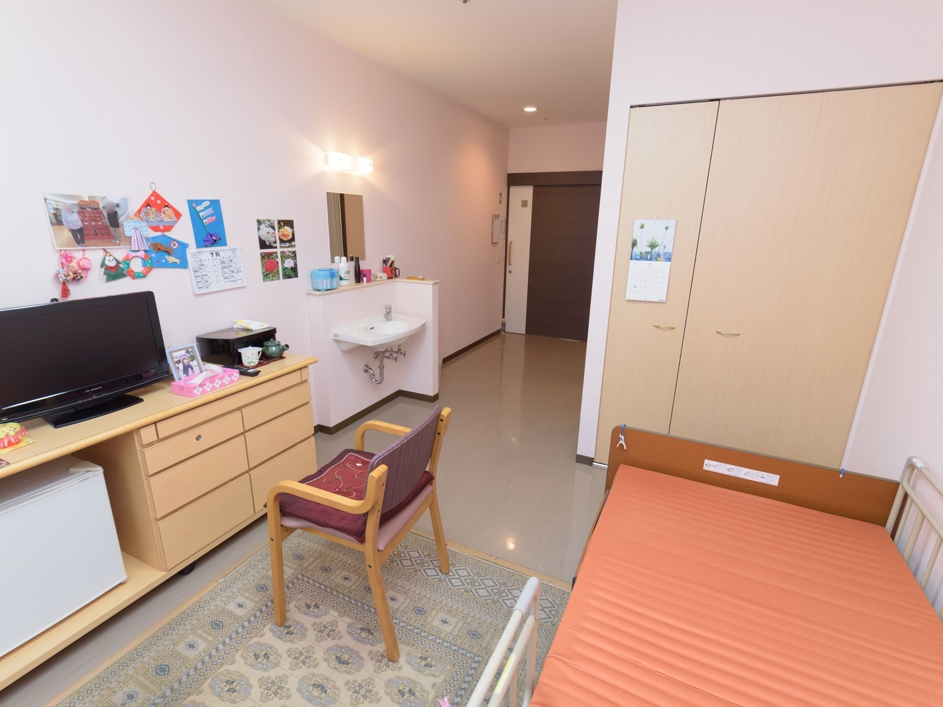 居室にはベッドや椅子、棚、テレビなどが置かれている。壁には写真などが貼られており、近くには洗面台が設置されている。