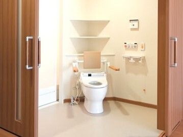とびらが２方向に開け閉めできるようになっている広いトイレ。便座には手すりと背もたれが付き、呼び出しボタンや操作ボンタンが壁に設置されている。