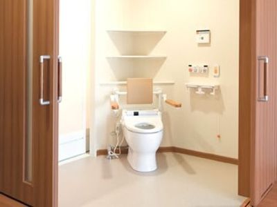 とびらが２方向に開け閉めできるようになっている広いトイレ。便座には手すりと背もたれが付き、呼び出しボタンや操作ボンタンが壁に設置されている。