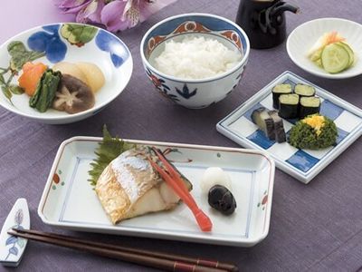 白いご飯に野菜の炊き合わせ、焼き魚に漬物の盛り合わせなどが、それぞれ和風の器に盛られ、箸置きには箸が置かれている。