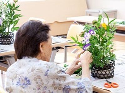 和風のフラワーアレンジメントを楽しむ高齢女性。真剣な表情でしている。新聞心広げられたテーブルの上にははさみも置かれている。