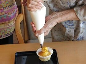 小さなココット型の焼き菓子の上にオレンジ色のクリームを絞り出している様子。クリームを持っているのは高齢者の手。