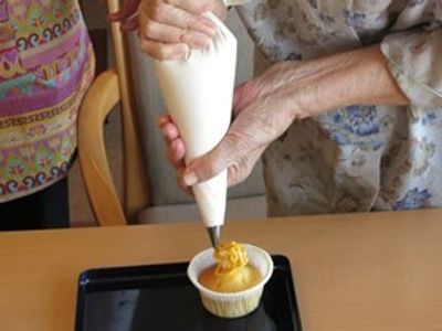小さなココット型の焼き菓子の上にオレンジ色のクリームを絞り出している様子。クリームを持っているのは高齢者の手。