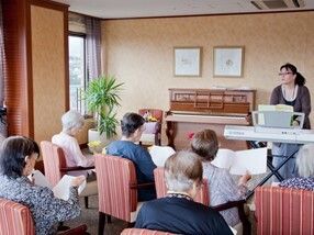 赤い椅子が並べられ、女性入居者が冊子のようなものを見ながら演奏を聞いている。演奏者は眼鏡をかけた女性で電子ピアノを弾いている。