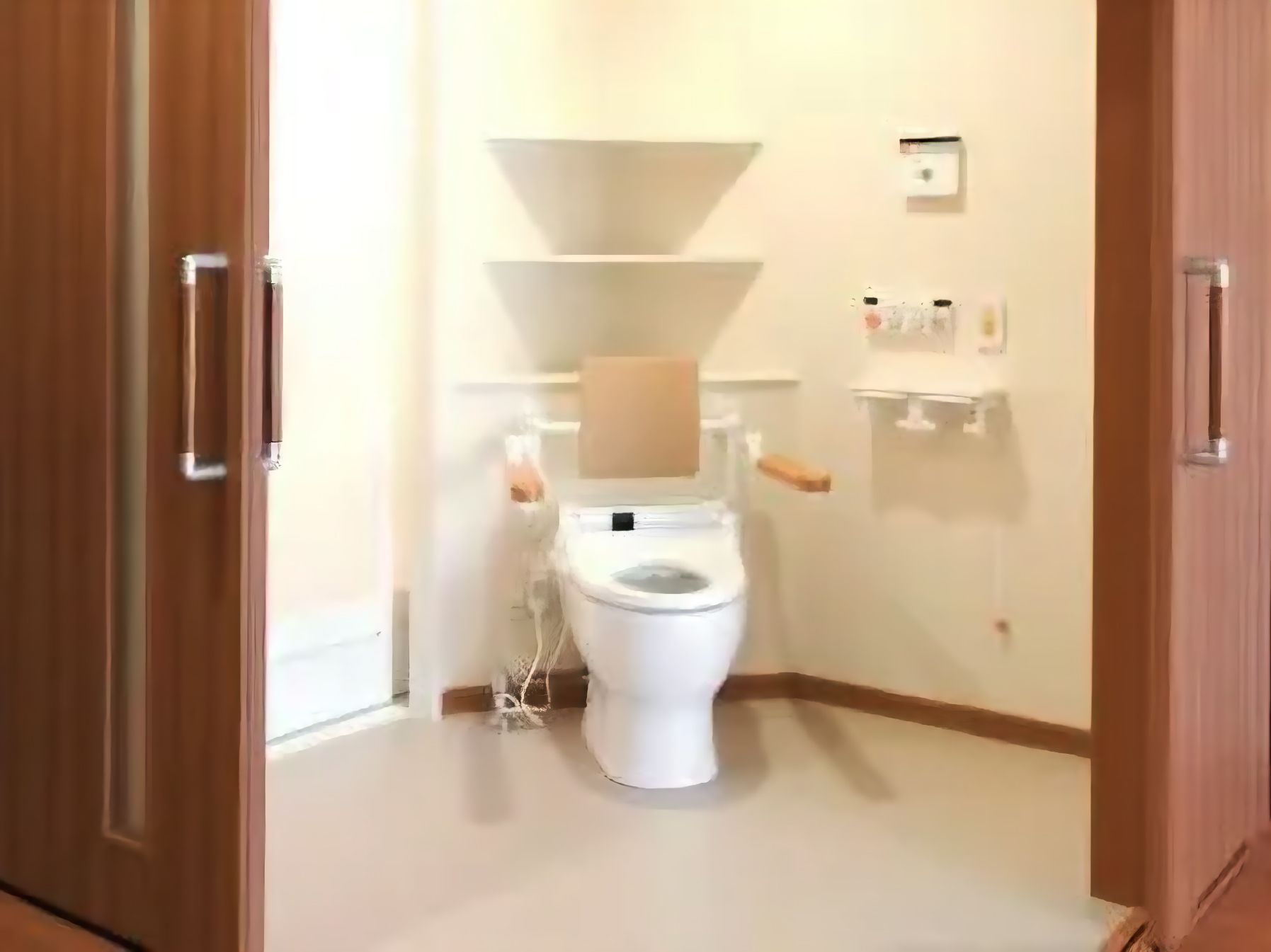 とびらが２方向に開け閉めできるようになっている広いトイレ。便座には手すりと背もたれが付き、呼び出しボタンや操作ボンタンが壁に設置されている。