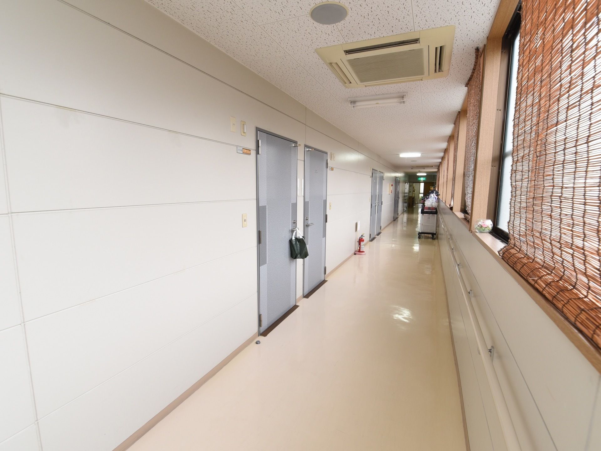 廊下には窓とグレーのドアが並んでいる。窓の下には、白い手すりが続いている。天井には、エアコンが設置してある。