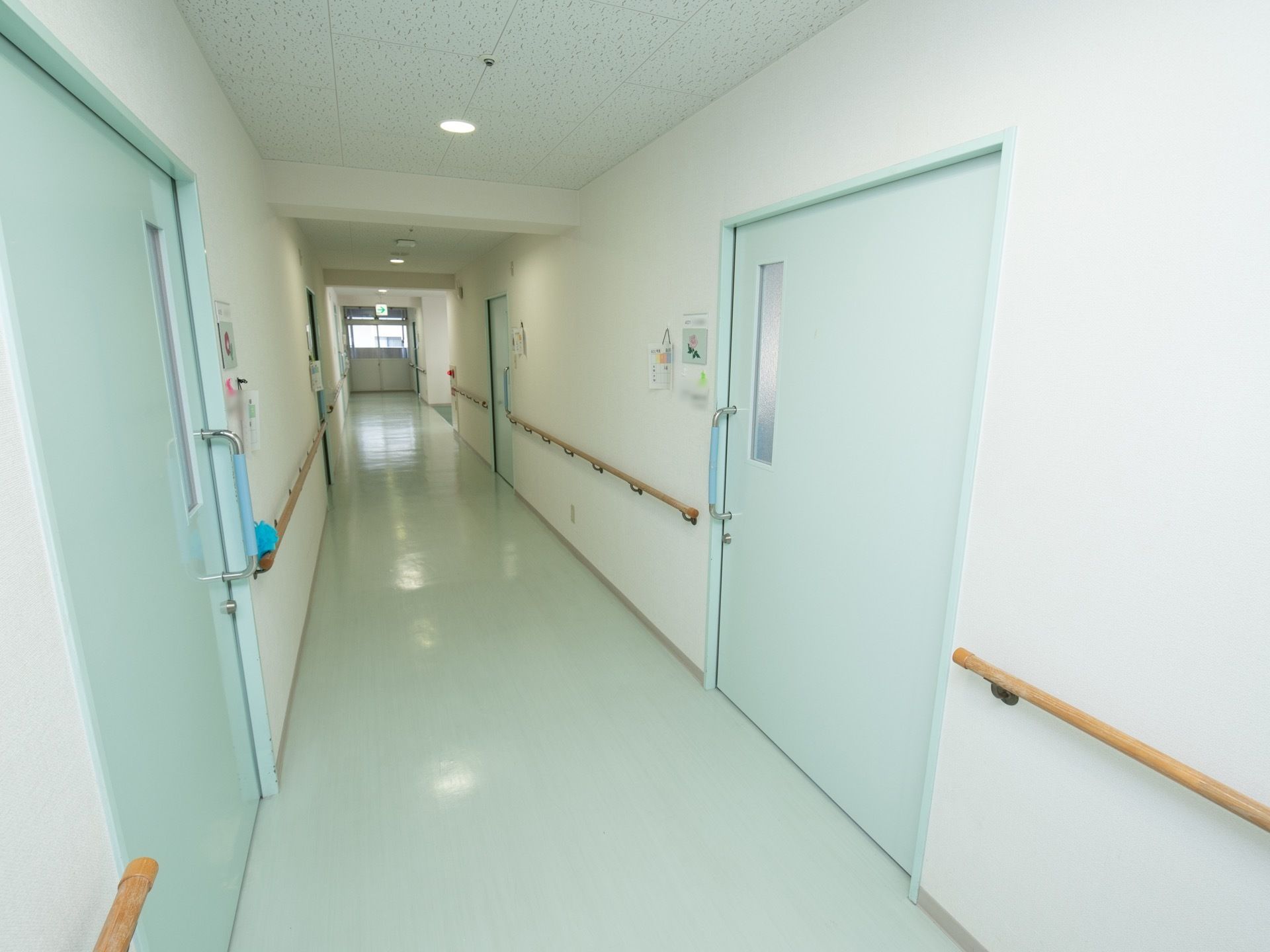 廊下の床面と居室入口の扉は、薄い水色で統一されている。壁には、丸い木の手すりが連続して設置されている。