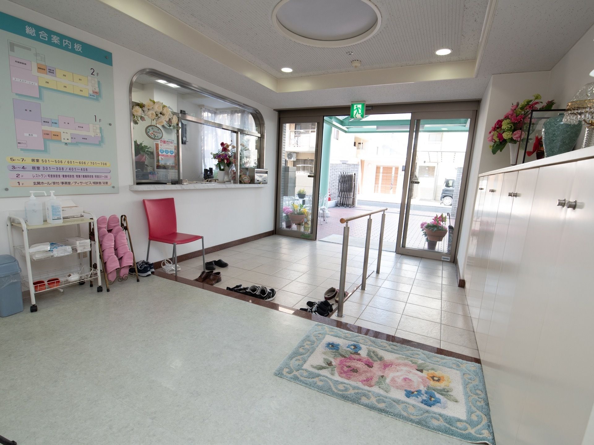 玄関の土間に、赤色の椅子が１つ置かれている。施設内の案内図の下に、来訪者様用のスリッパのラックがある。