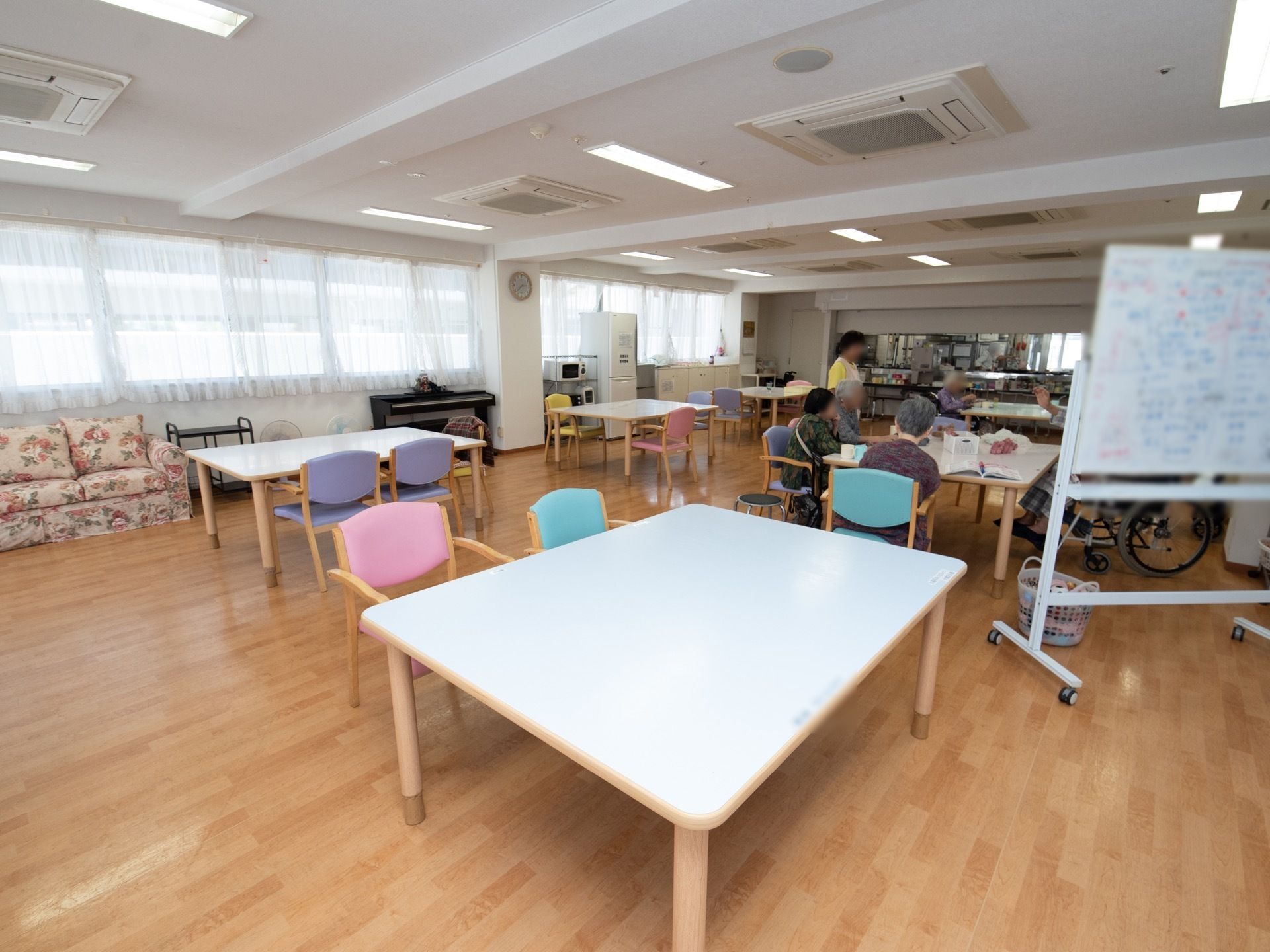 食堂に白色の天板のテーブルが６台置かれている。入居者様が何人か集まっていて、その近くに、１人のスタッフが立っている。