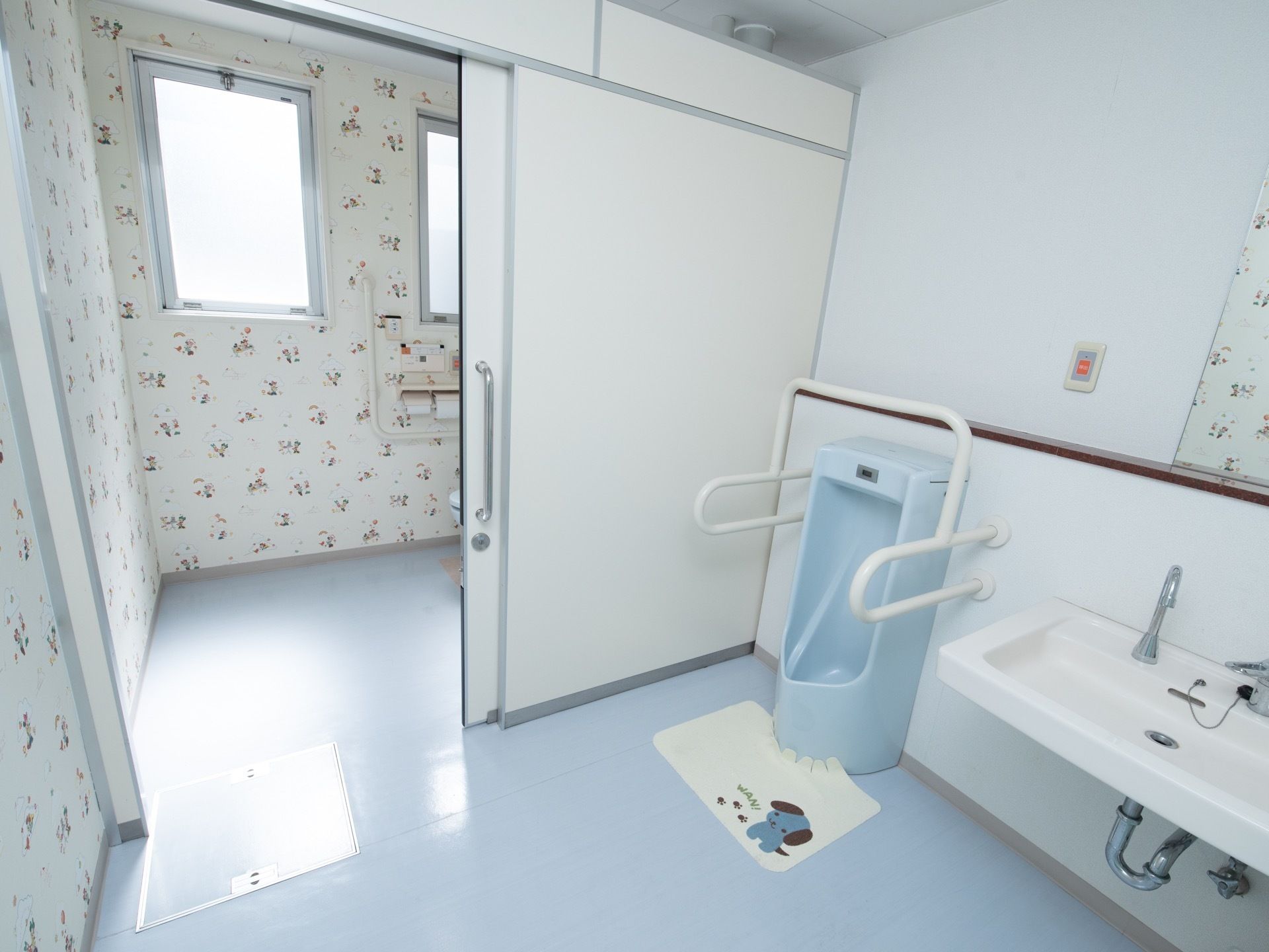 トイレの壁に、かわいい模様の壁紙が貼られている。男性用便器の手前には、尿はね防止のマットが敷かれている。