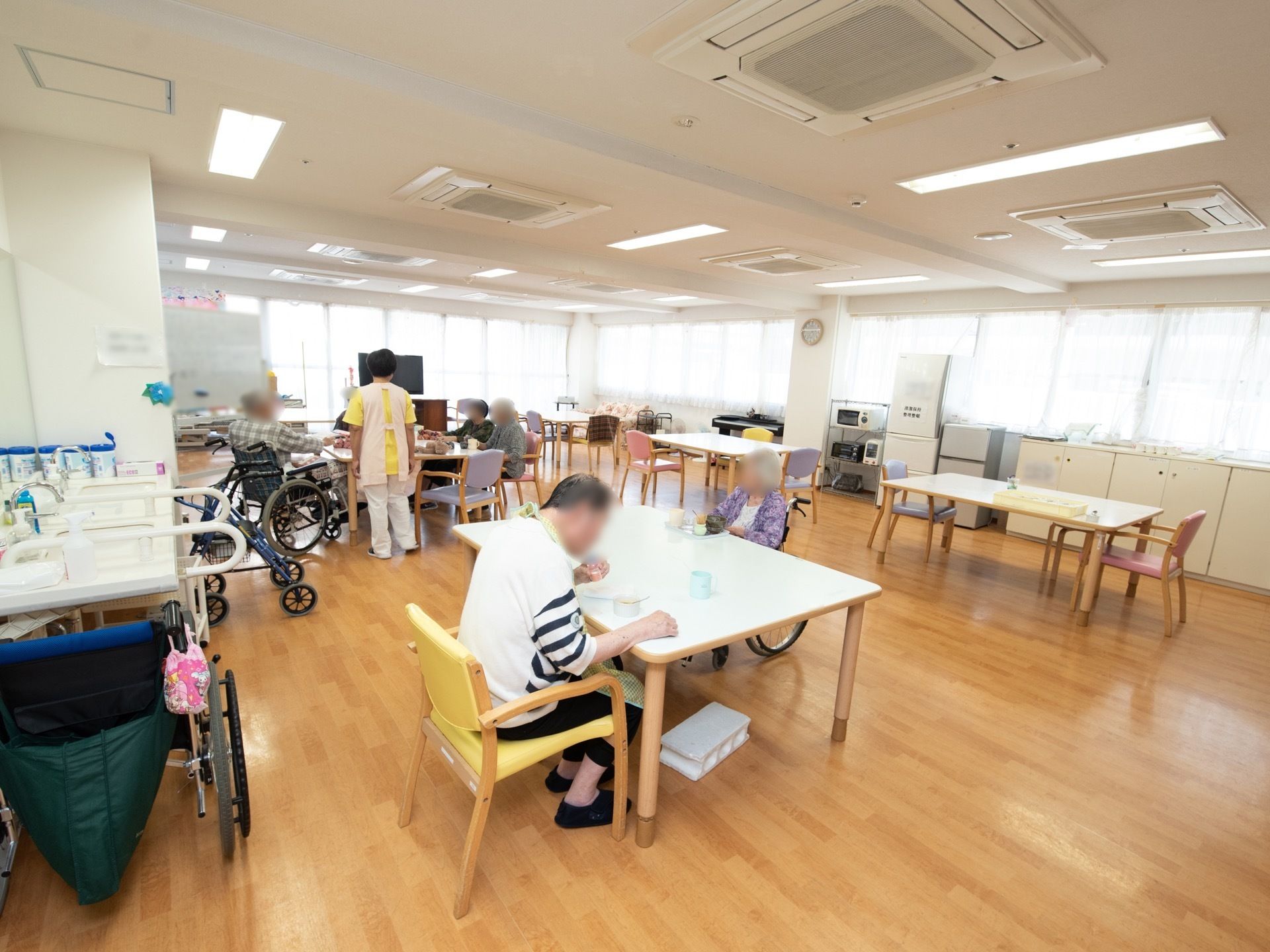 食堂に入居者様が何人か集まっており、その近くに女性スタッフが１人立っている。テーブルの下には、足置きが置かれている。