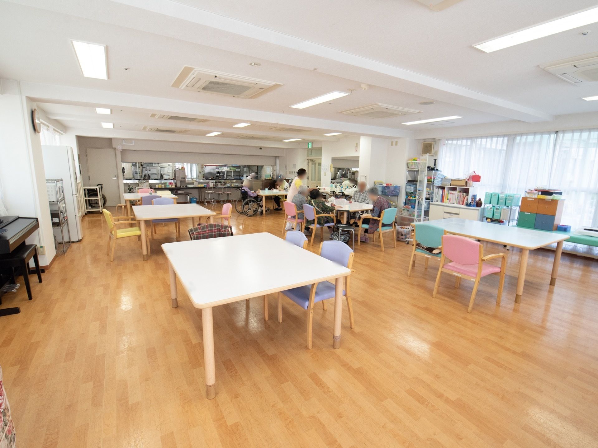 食堂は、大きな窓からの採光や多くある照明で明るい。入居者様が集まっているテーブルの横に、女性スタッフが立っている。
